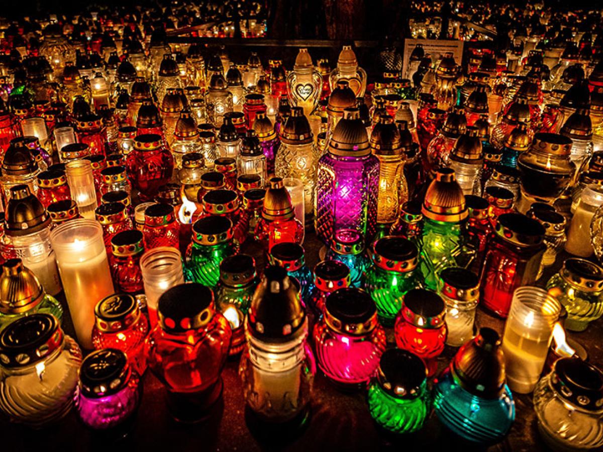 Dozens of candles in cloured jars and holders litter the ground of a cemetry.