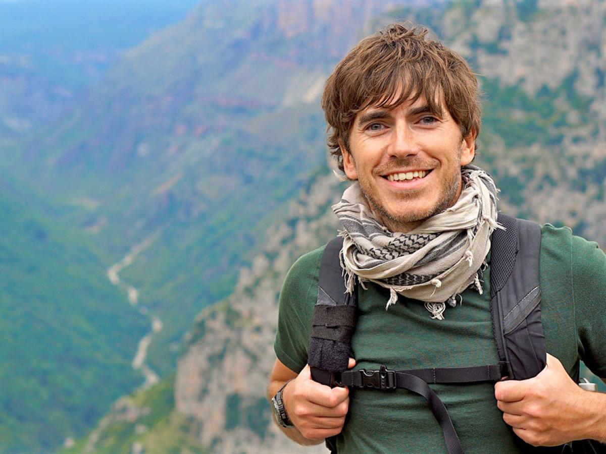 An enthusiastic hiker stands in front of a view down a valley, smiling and holding his backpack straps.