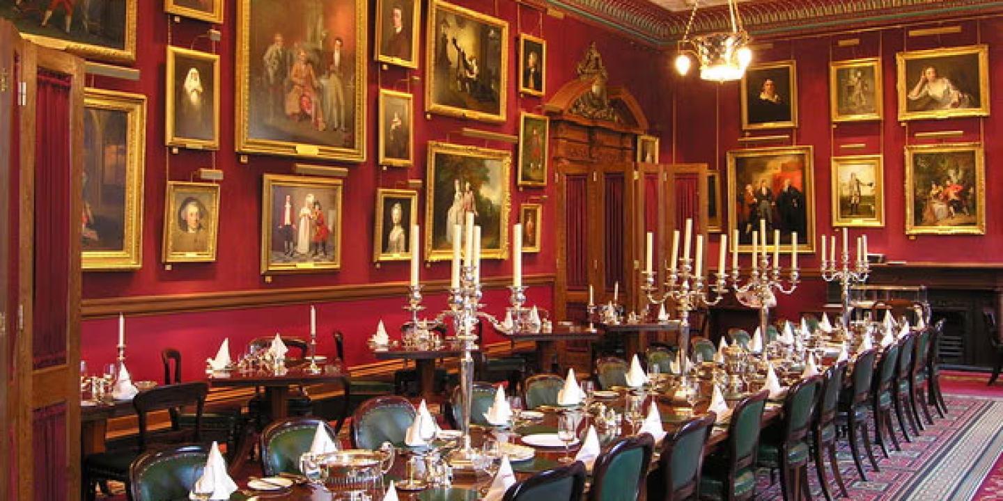 A grand dinner table set for a meal sits within a large room with paintings on the wall.