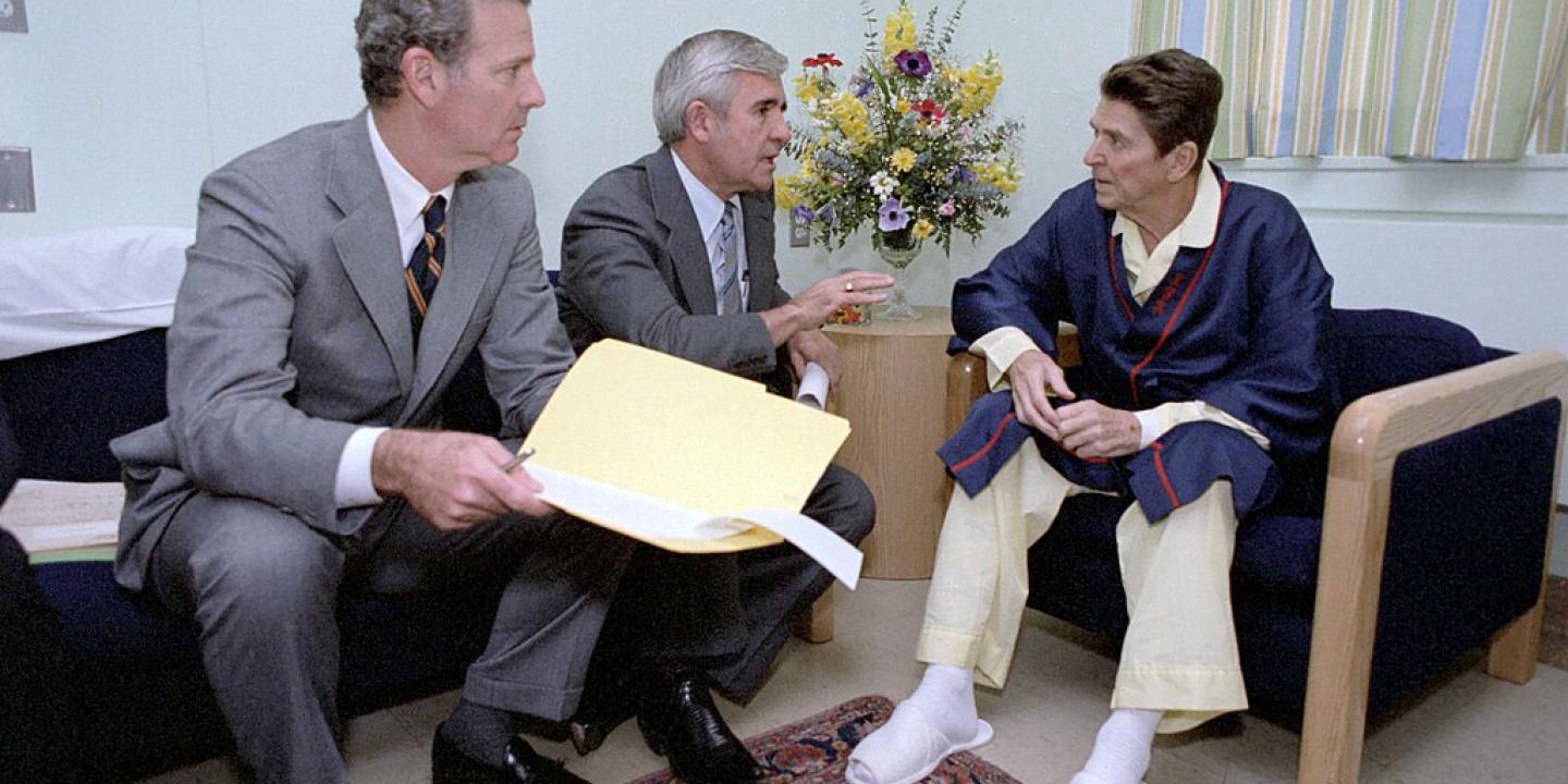 Two men in seats adress a pyjamap-clad Ronald Regan.