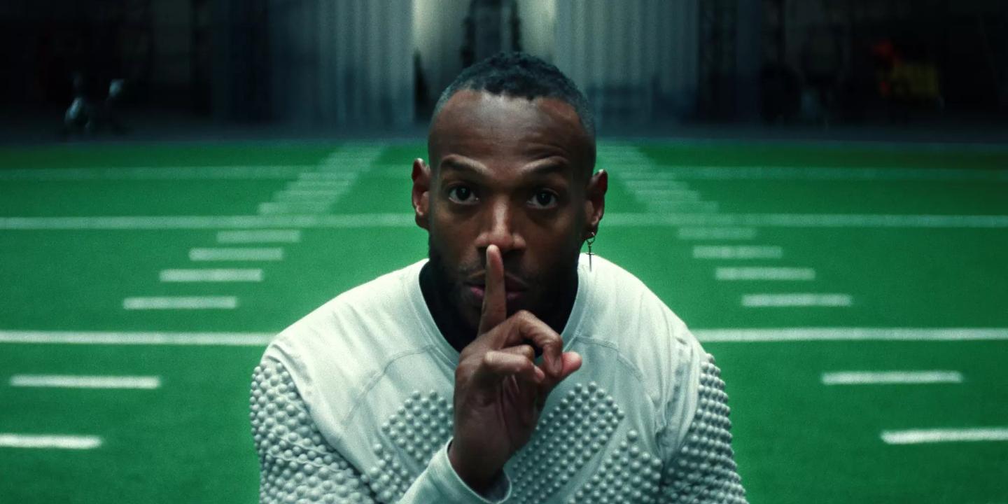 A player holding a finger to his lips stands in front of an indoor American football pitch.