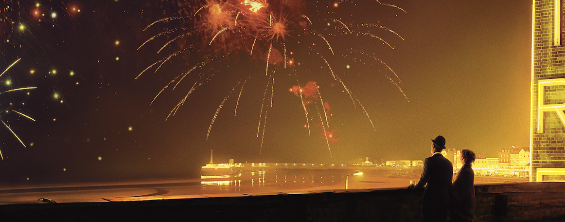 A couple stand on a seafront watching fireworks explode over a beach and pier. Credut