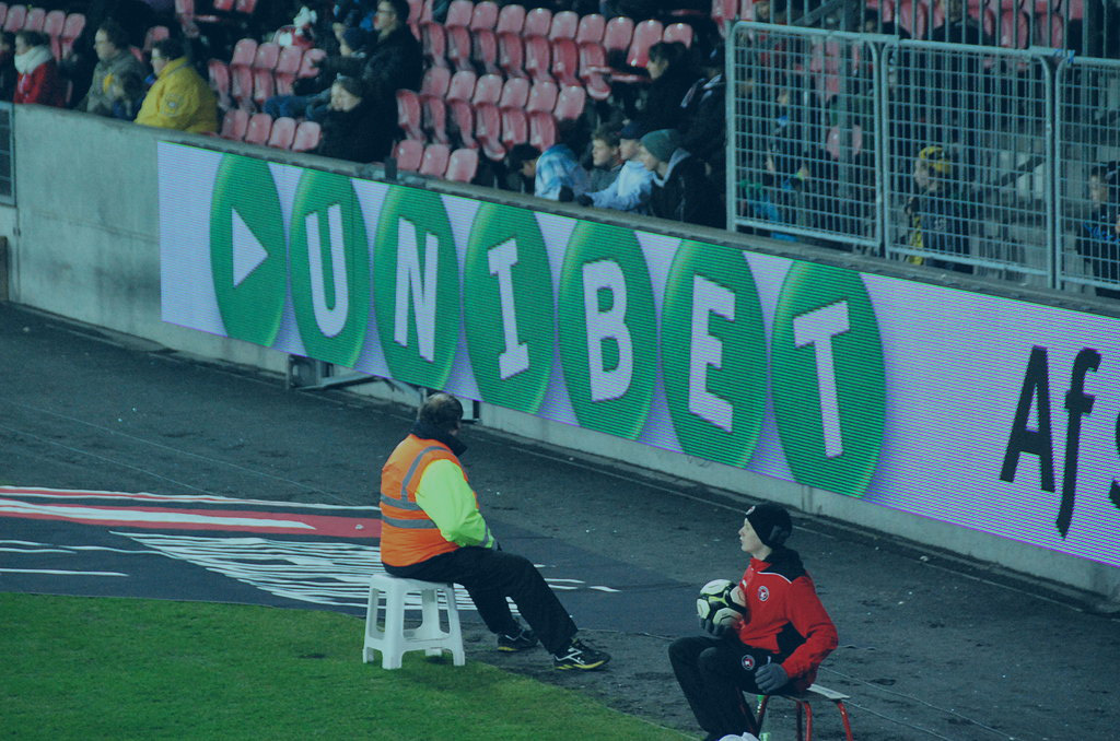The edge of a football pitch showing an advertising hoarding with a betting brand name on it.