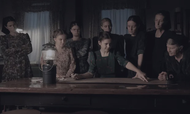 a group of women stand and sit around a table lit by a gas lamp.