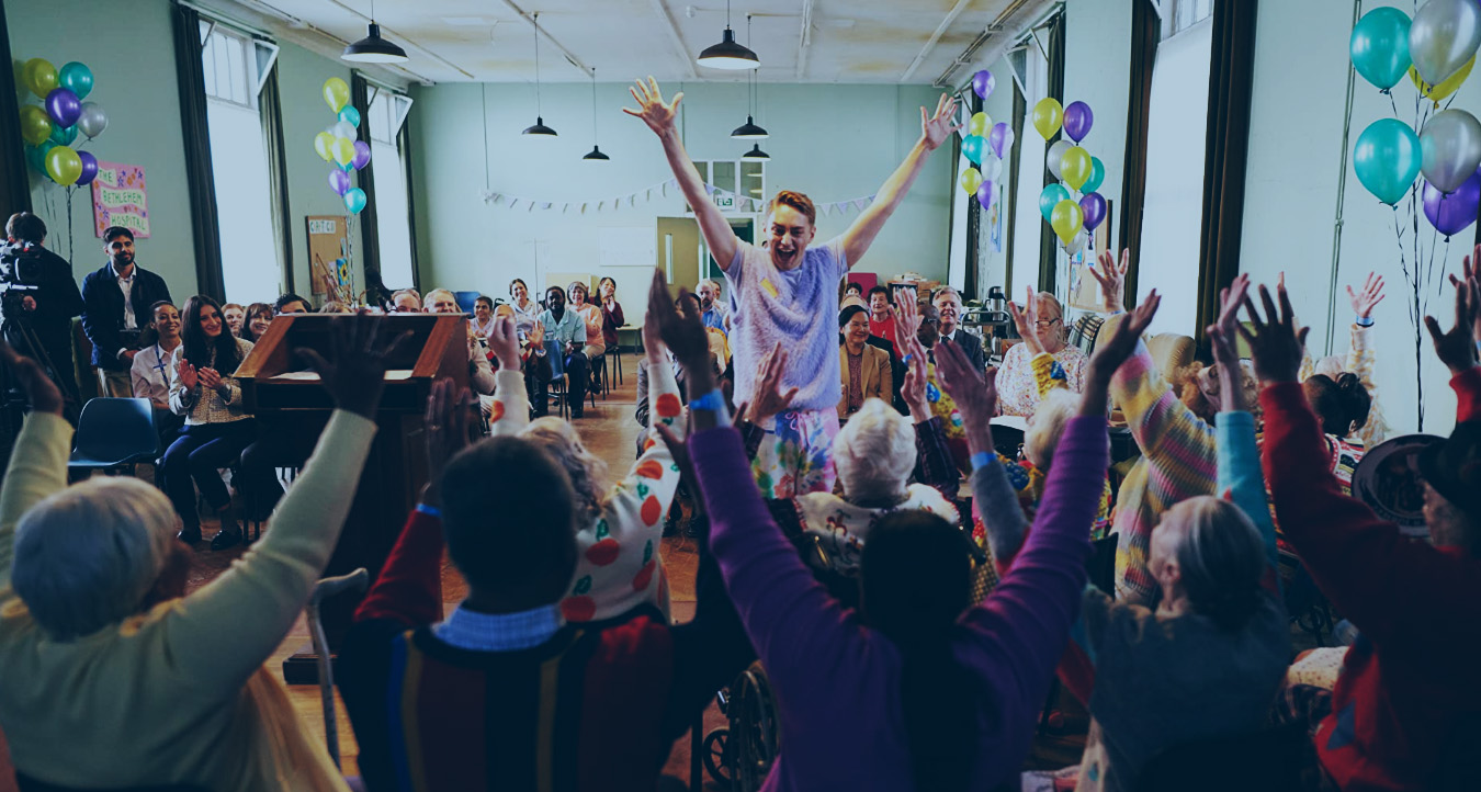 In a hall decorated for a celebration a person stands in front of a seated group, all have their arms raised in celebration.