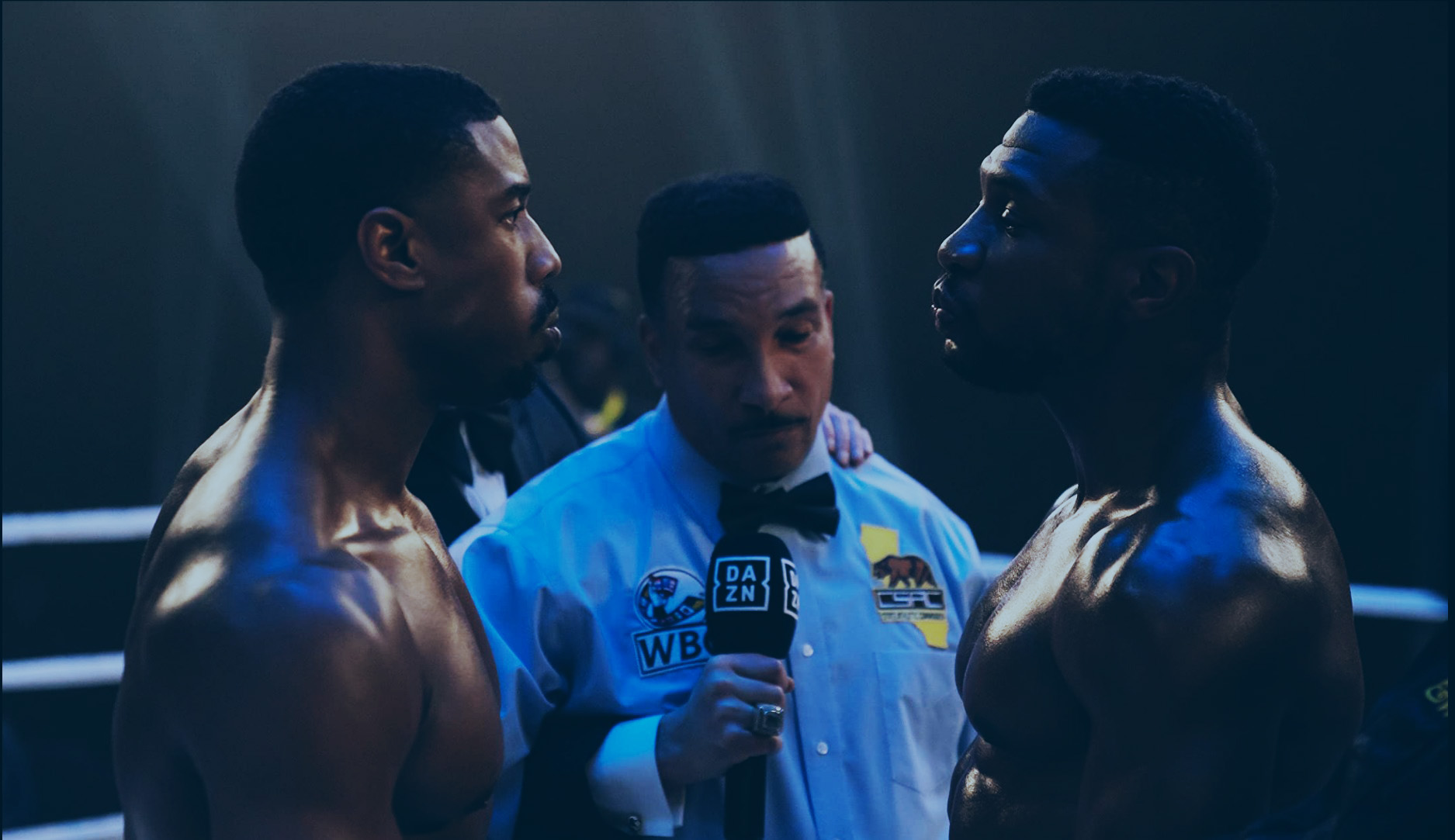 In a boxing ring, an umpire stands between two boxers who stare hard at each other.