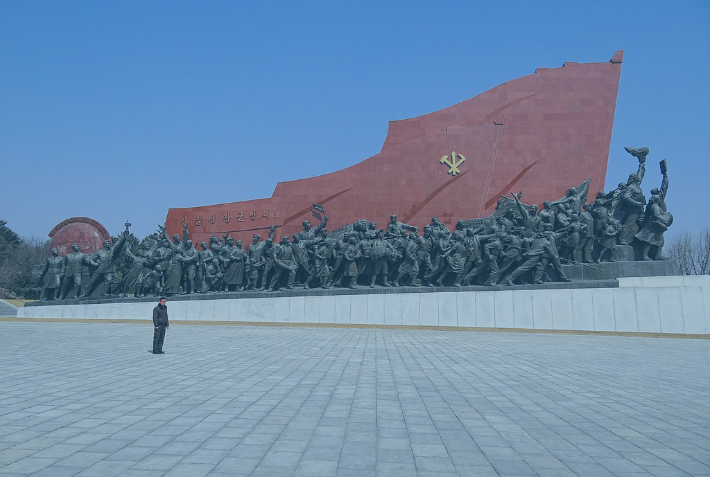 A huge communist monument consists of a red flag wall rising from left to right over a column of statues.