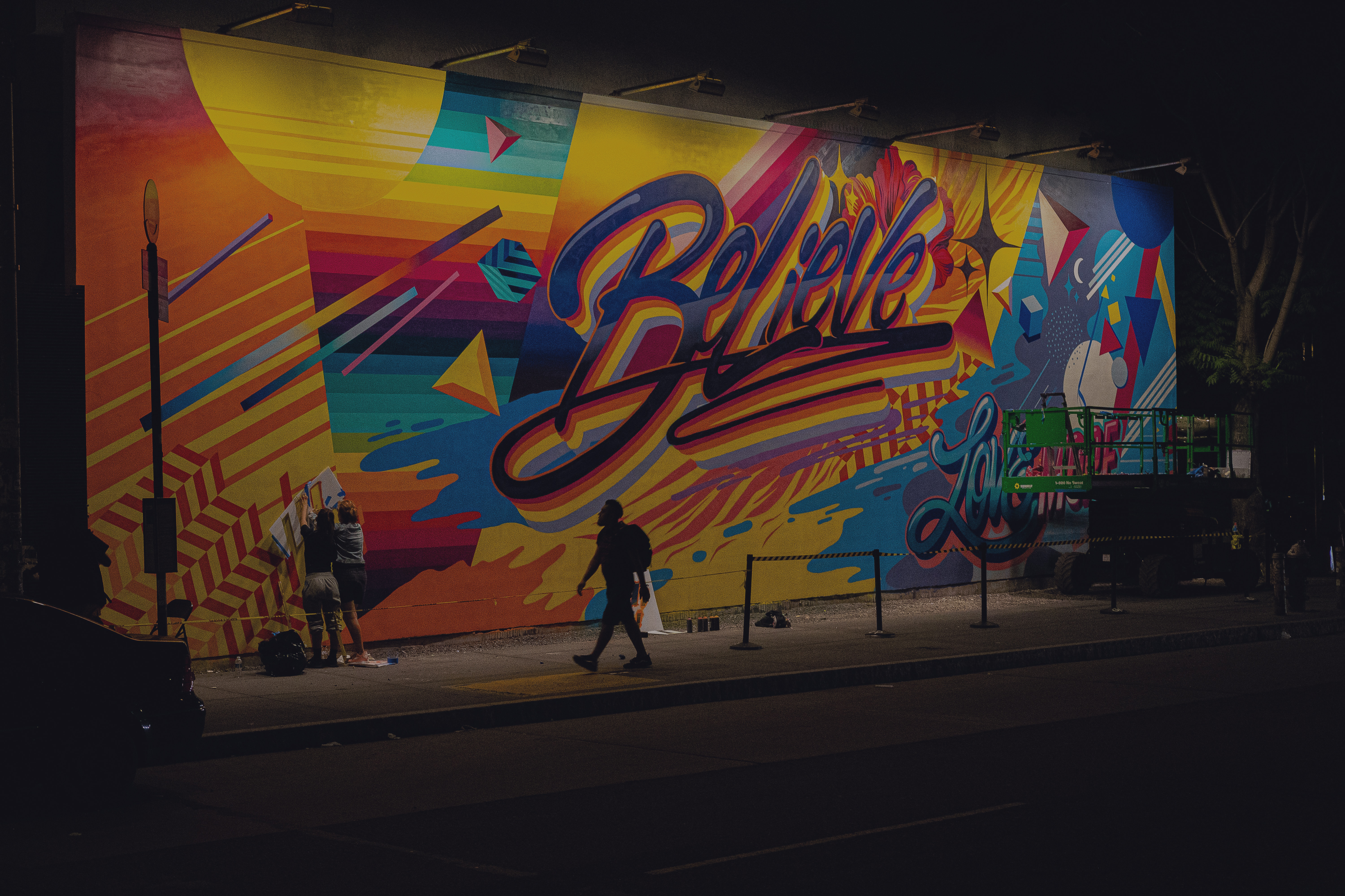 A person walks past a multi-coloured wall of graffiti with the word 'believe' in the centre of it.