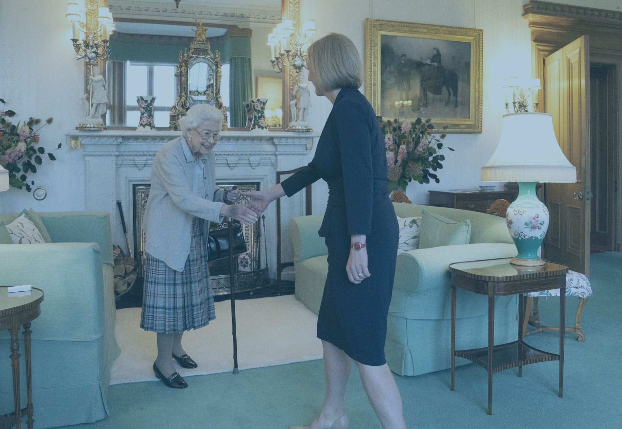 A frail old lady, the late Queen, rises from a sofa to shake hands with an approaching woman.