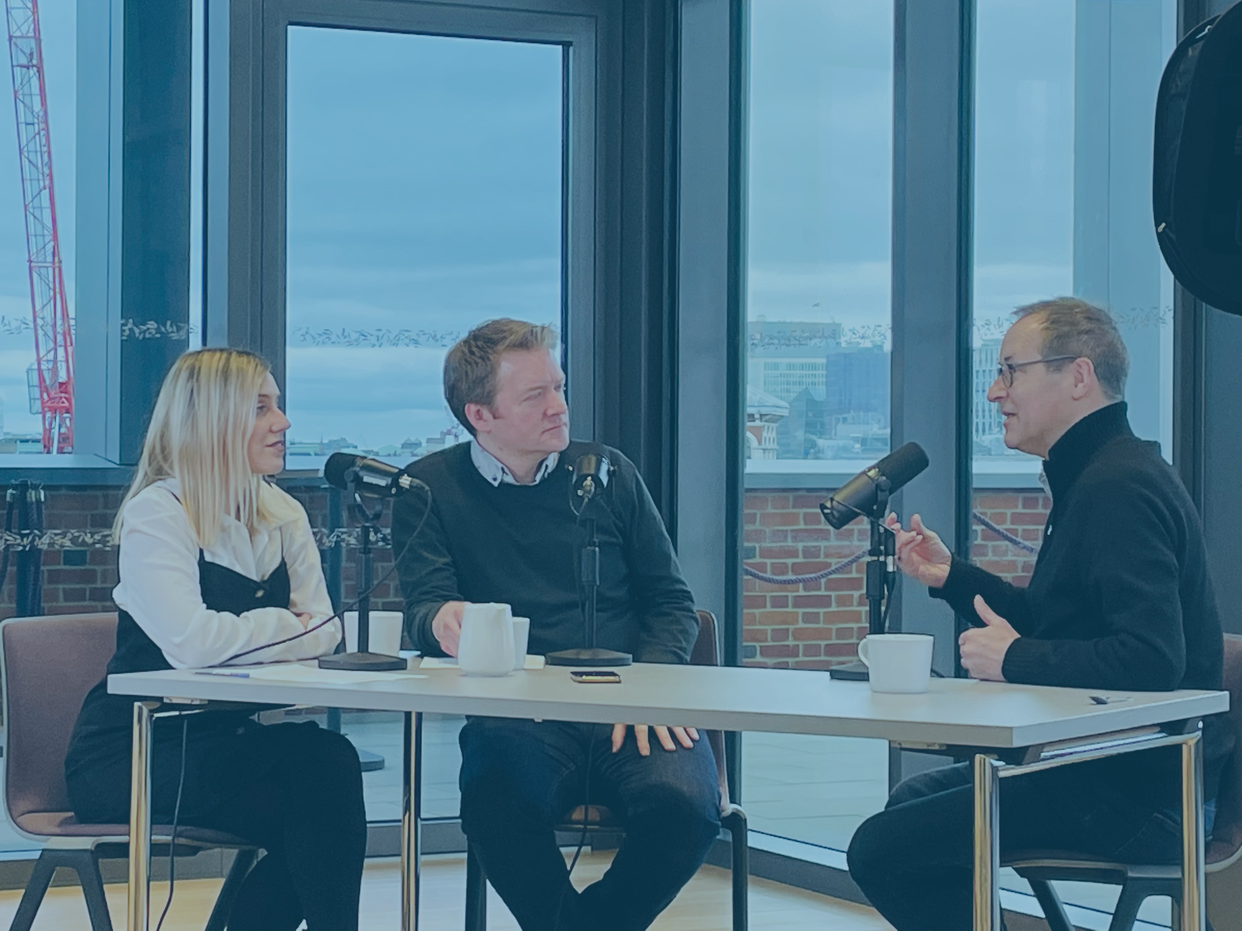 Three people sit around a table recording in front of microphones, one gestures with their hand
