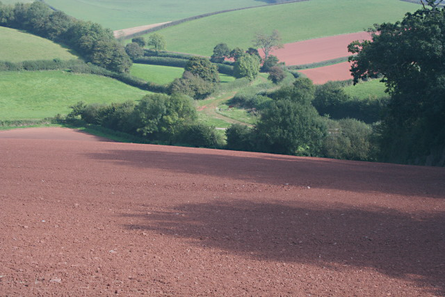 A ploughed field of red soil is in the foreground, sloping down into a valley with a track and green fields beyond