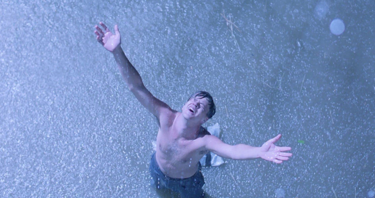 An man stands in the rain, topless, with face and arms raised in celebration.