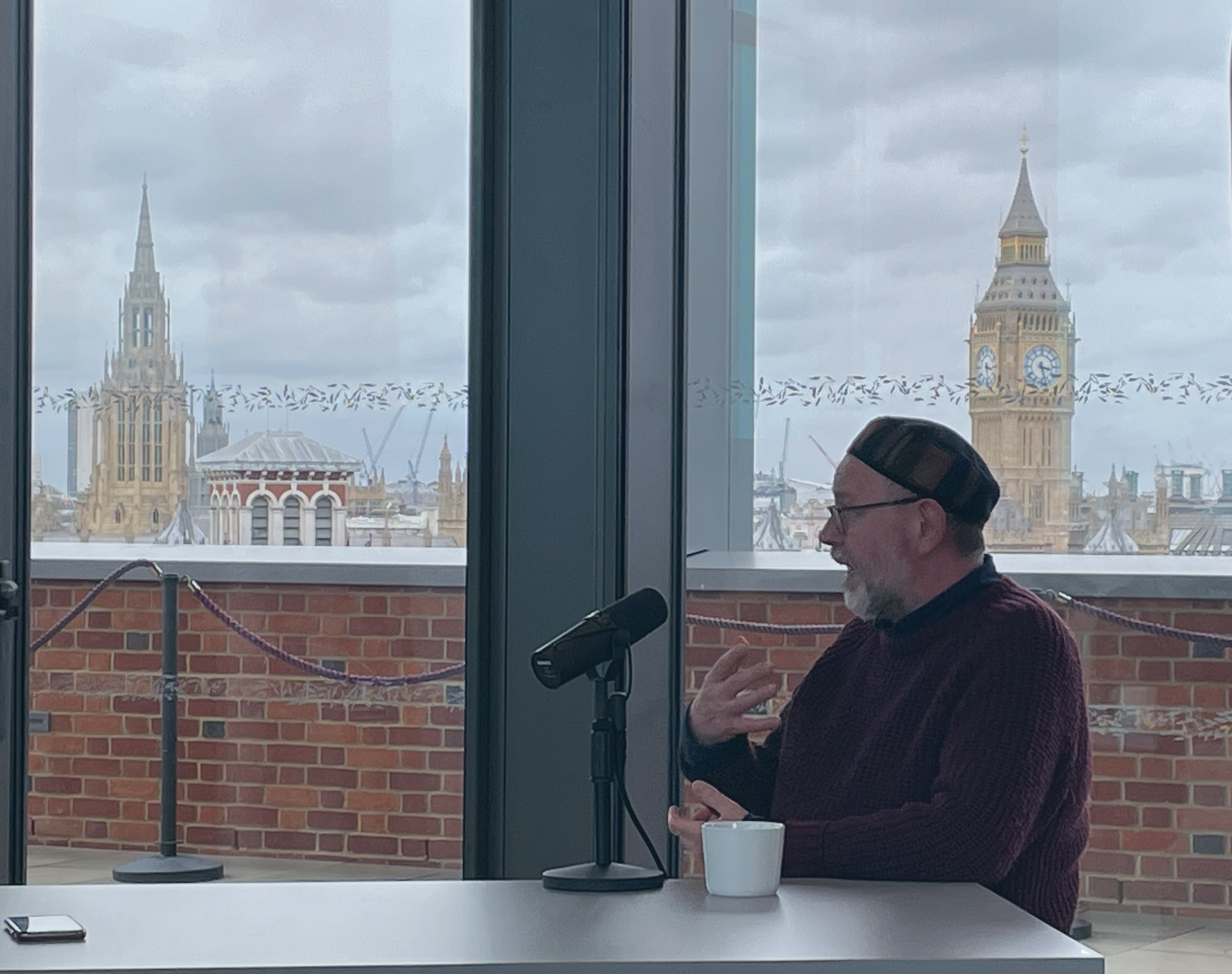 A man sits and speaks into a microphone beside him on a table, while gesturing with his hand. Behind, Big Ben is on the skyline.
