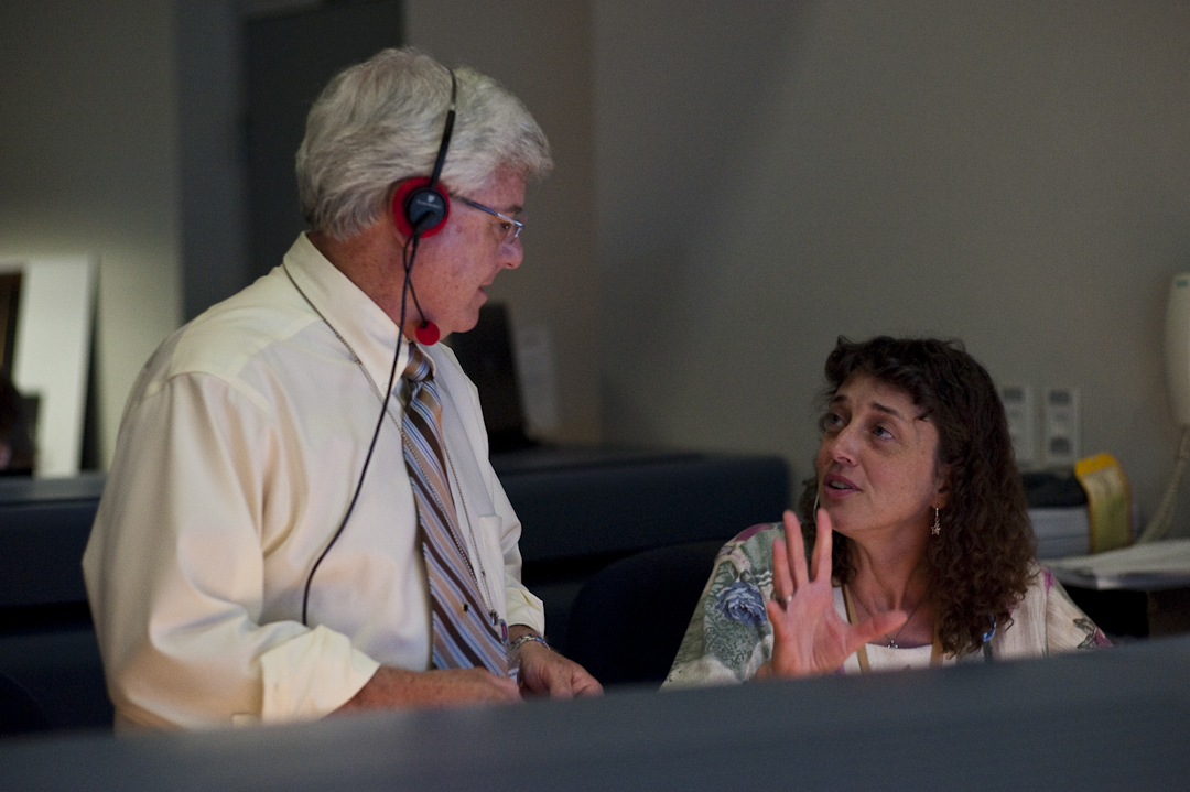 Two scientists talk to each other at console in a space mission control room.