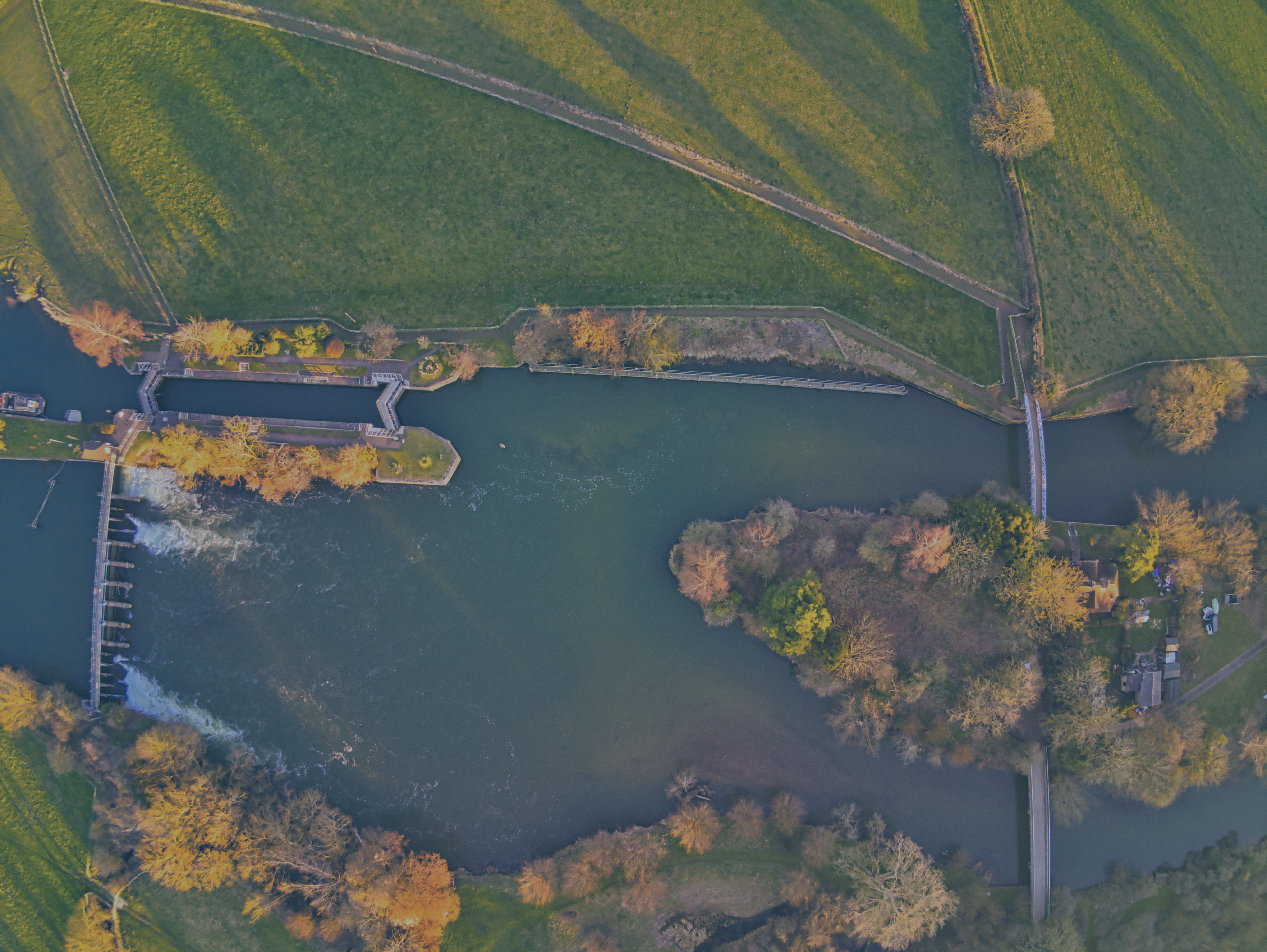 A bird's eye view of a river, with a lock and weir to the left and an island with two bridges to the right.