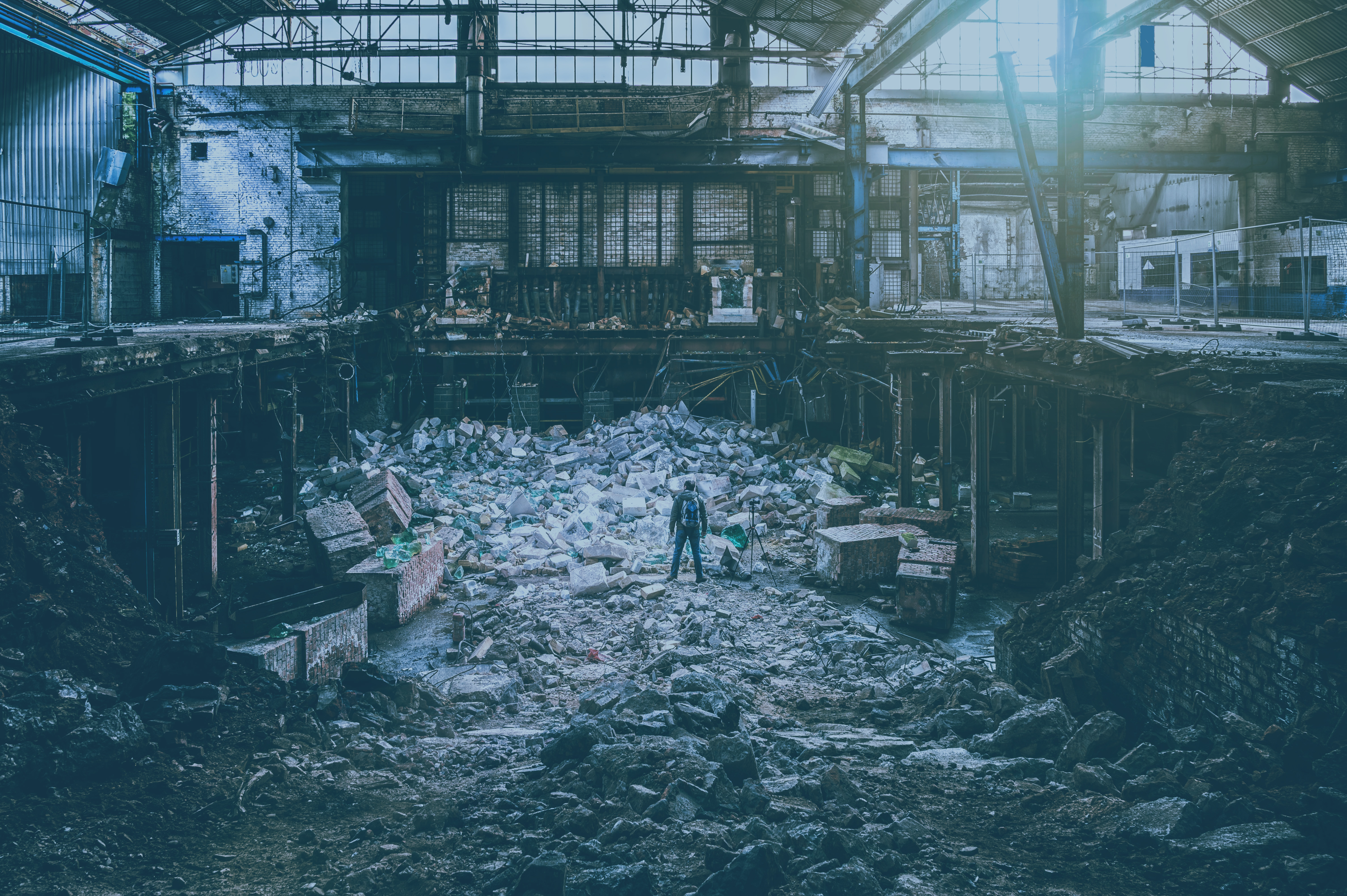 A photographer, standing next to a tripod, atop a pile of rubble is a destroyed factory.