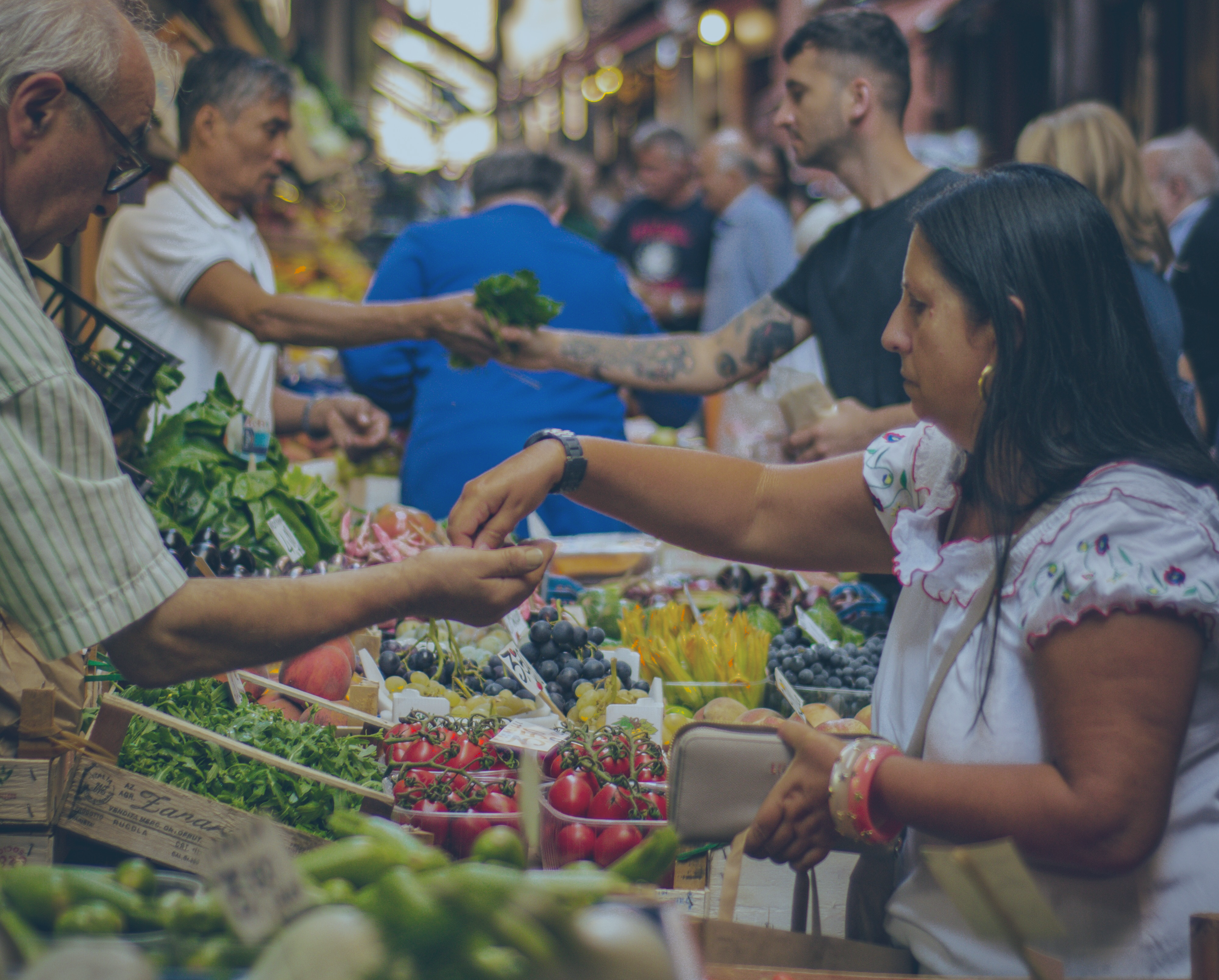 Marketer stall traders and customers reach out arms and hands to exchange fruit and money above piles of produce.