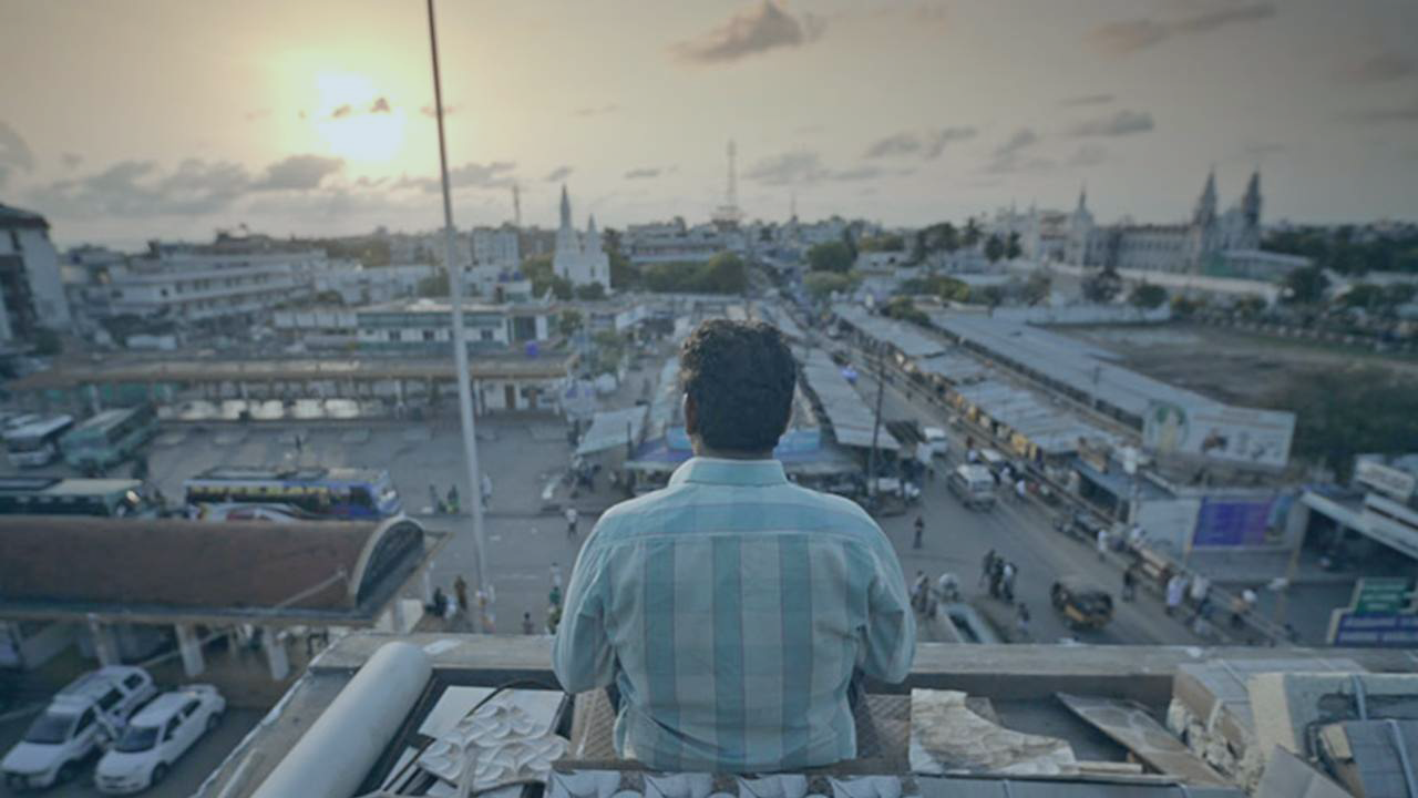 A man sits on the top of a building and looks out over the view of an Indian city as the sun sets.