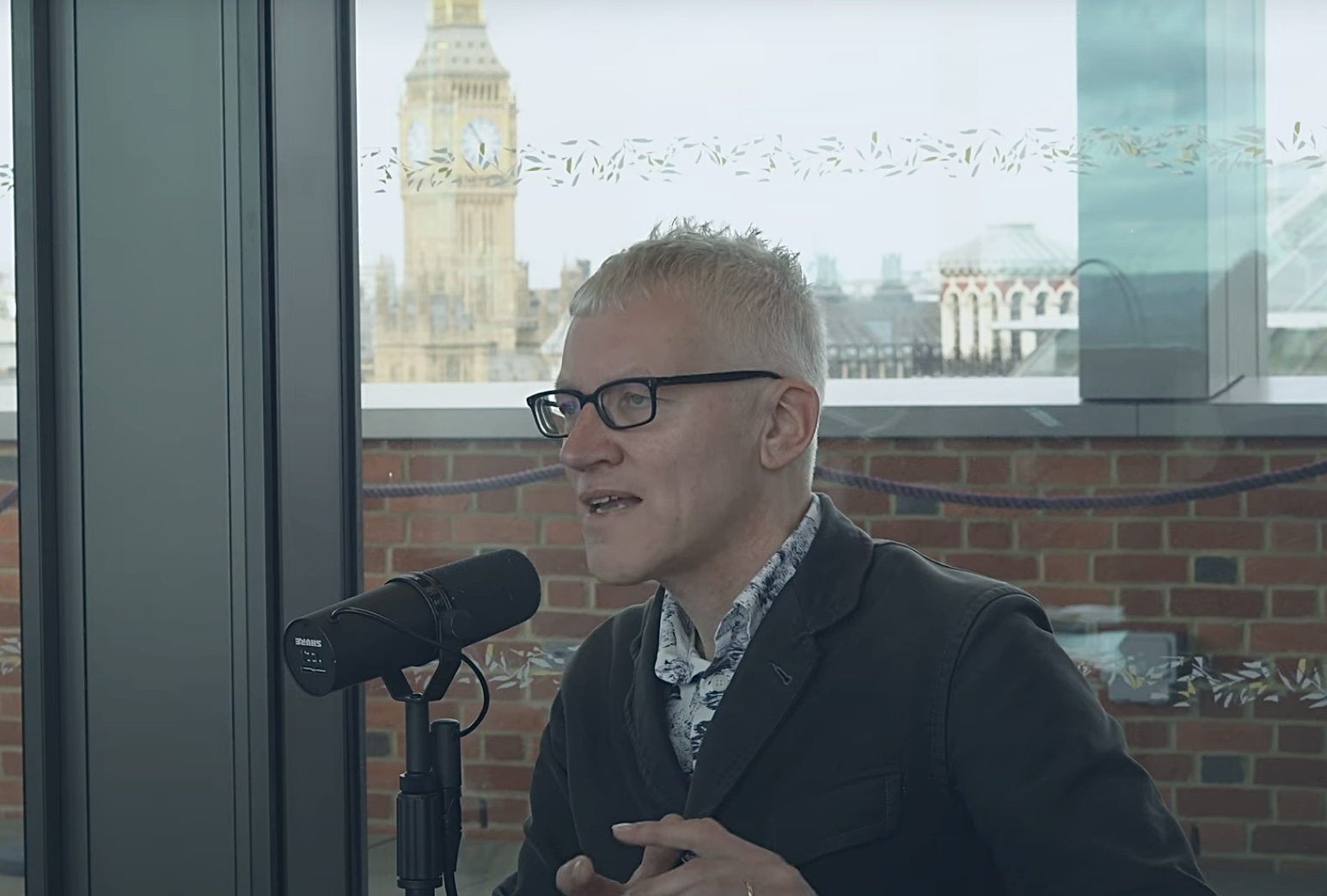 A man sits at a table speaking into a microphone but looks into the distance. Behind him, through the window and beyond a wall is Big Ben