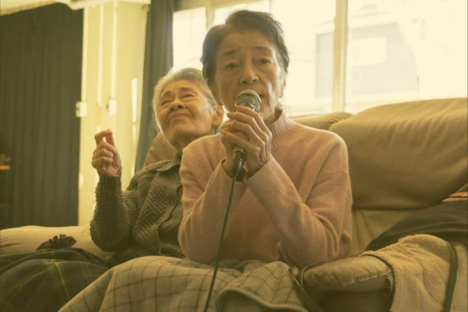 In a retirement home, a older person sings karaoke while the person behind waves a hand.