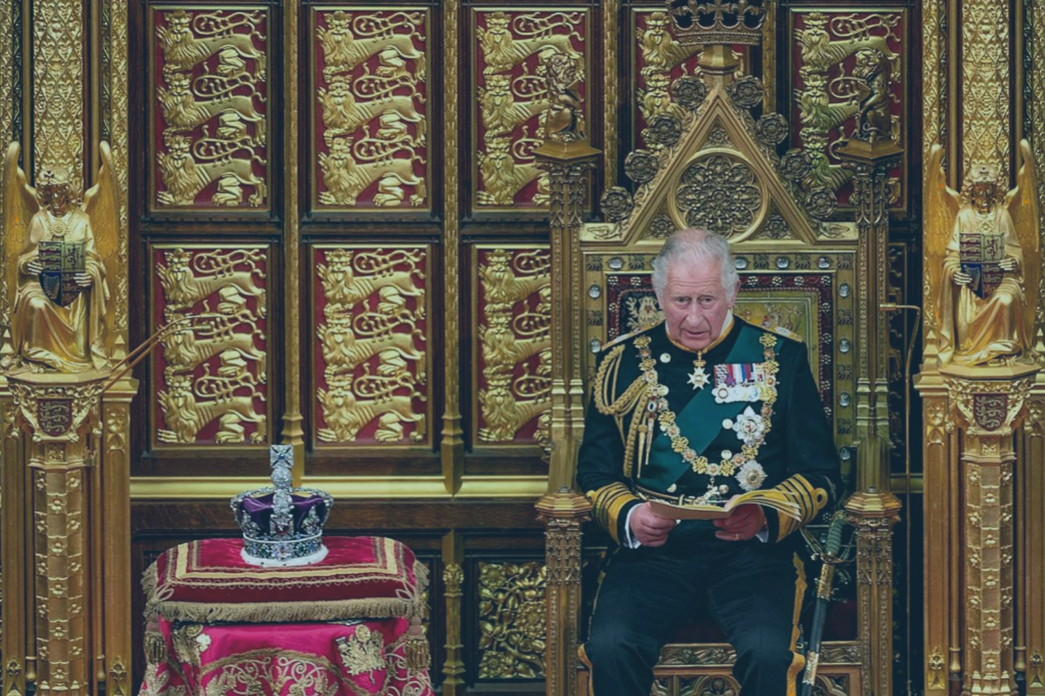 A uniformed Prince Charles sits on a throne reading a speech, beside a crown resting on a cushion
