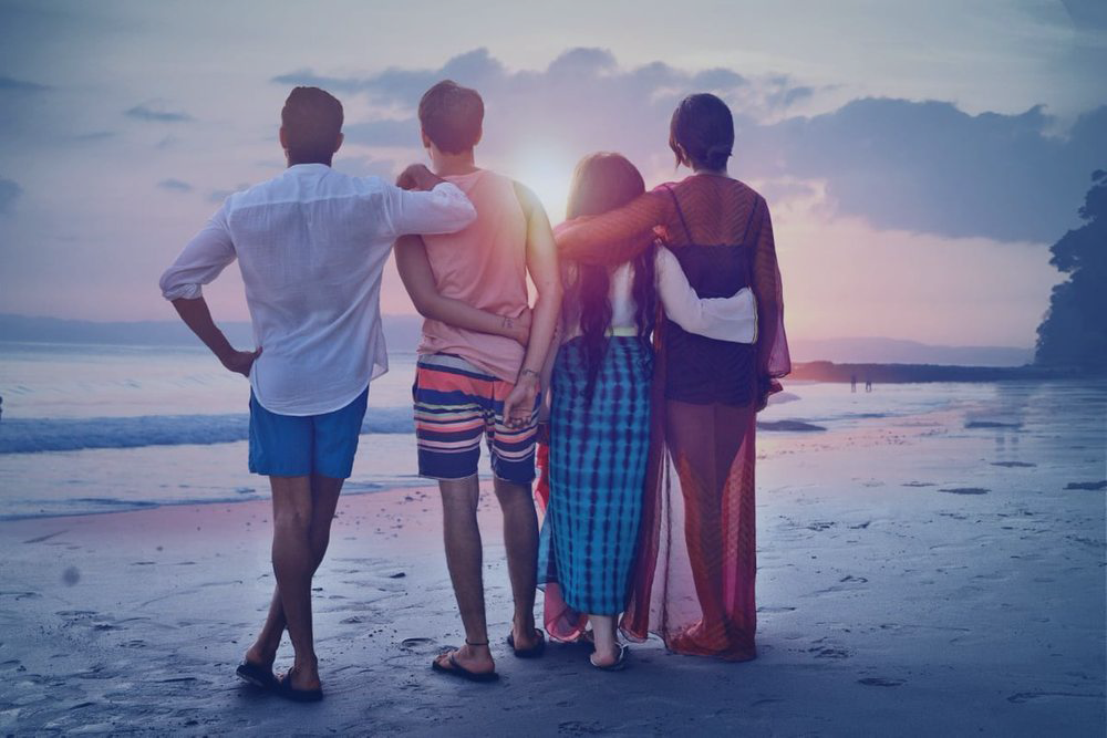 Parents and teenagers with their arms around each other stand on a beach and stare out to sea.