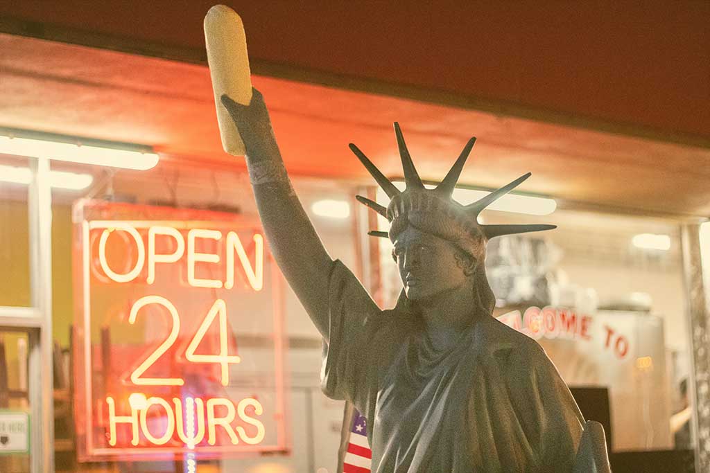 A copy of the Statue of Liberty, holding a stick of bread, stands outside a shop window displaying an 'Open 24 Hours' signs.