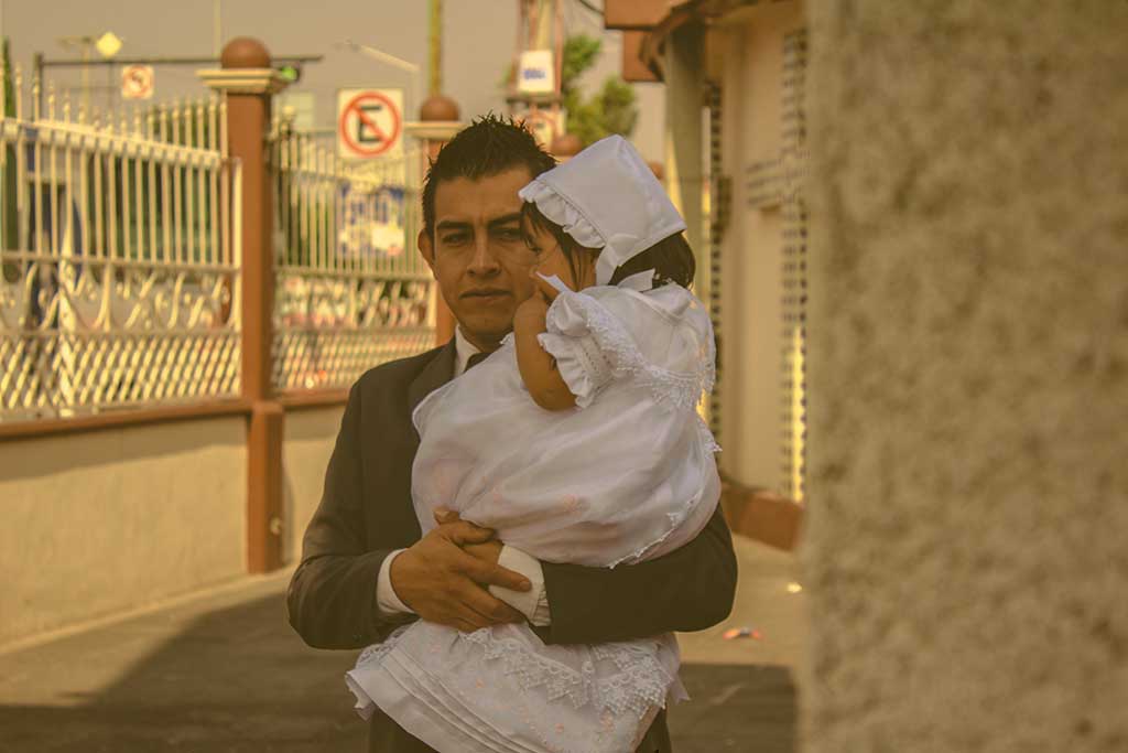 A father wearing a suit carries his child who is dressed for a christening in white.