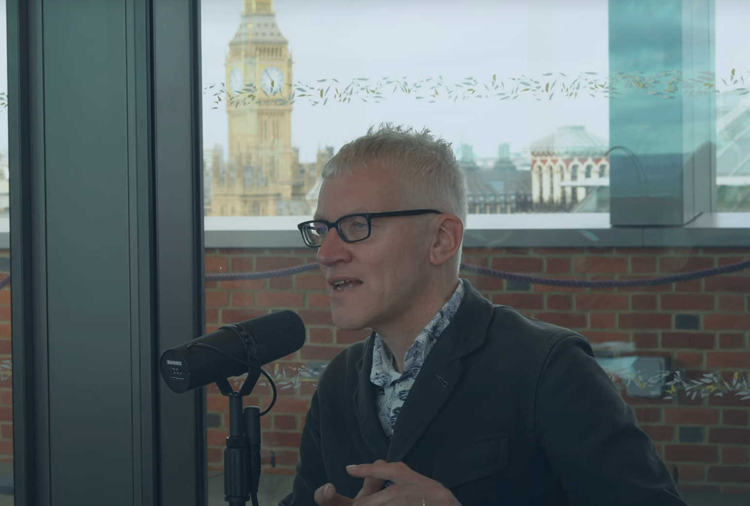 A man sits at a table speaking into a microphone but looks into the distance. Behind him, through the window and beyond a wall is Big Ben
