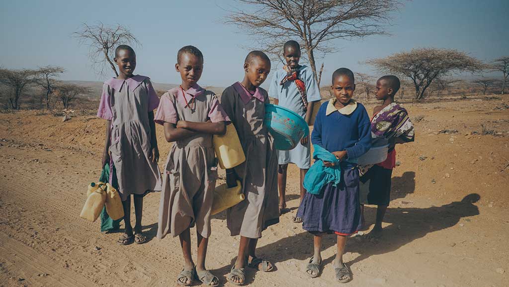 Girls stand in the northern Kenyan scrubland holding water bottles.