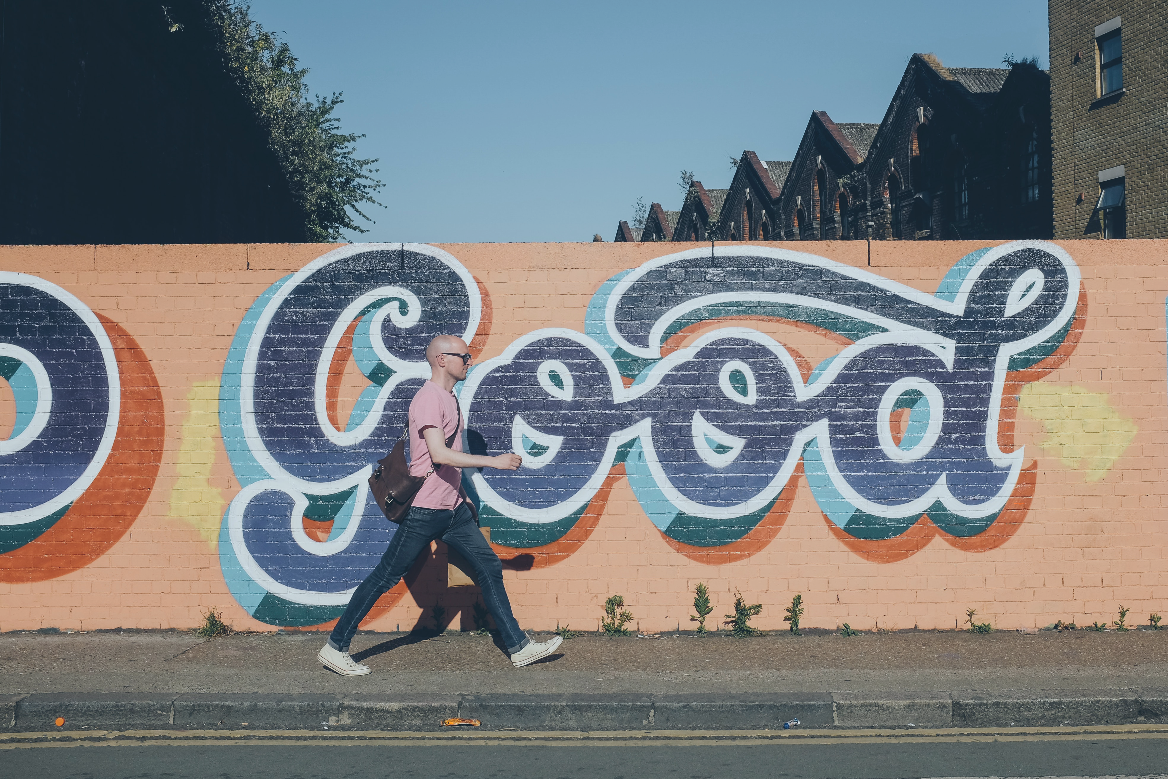 A man walks along a street past a orange wall with a huge 'Good' written in cursive script on it.