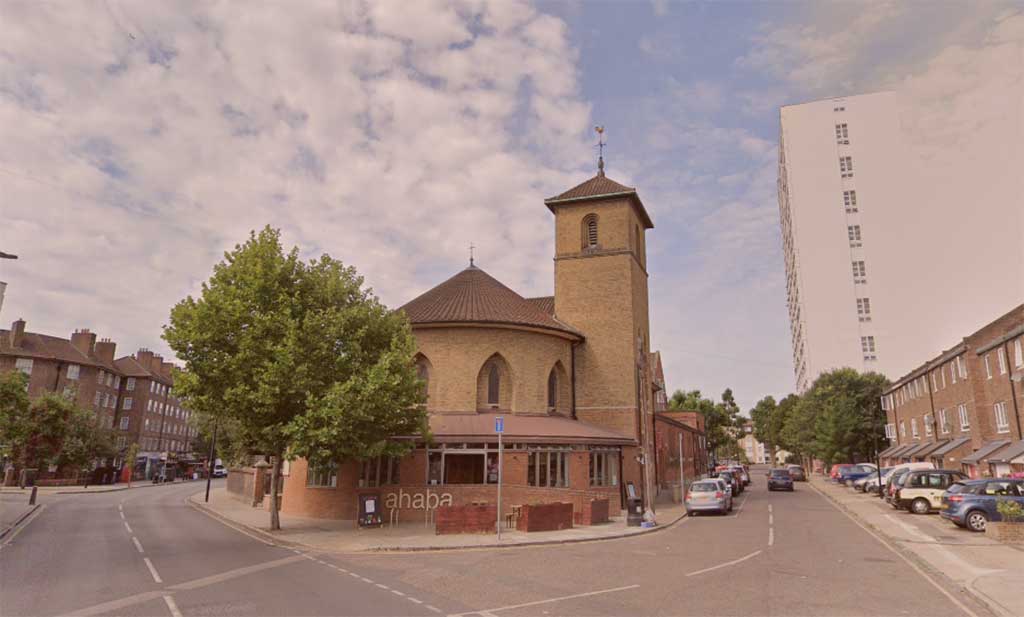 A neo-Romanesque church sits at the acute corner of two roads. To its side a tower block rises over a row of low-rise flats.