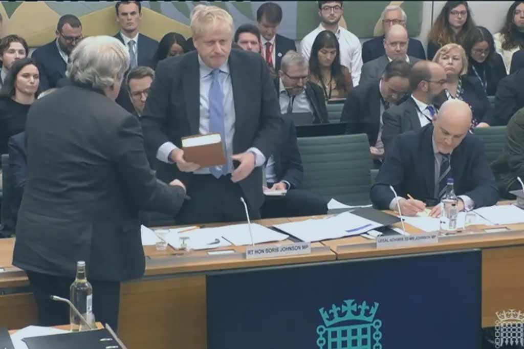 A politican stands holding a bible, in front of a committee room table. Behind him an audience waits expectantly