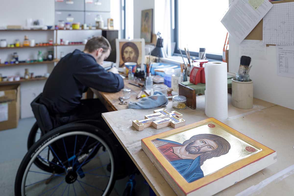 A monk in a wheekchair works on an icon in an art studio. In the foreground is a completed icon. 