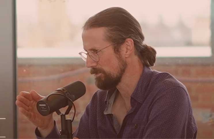 A sitting man speaks into a microphone will gesturing with one hand