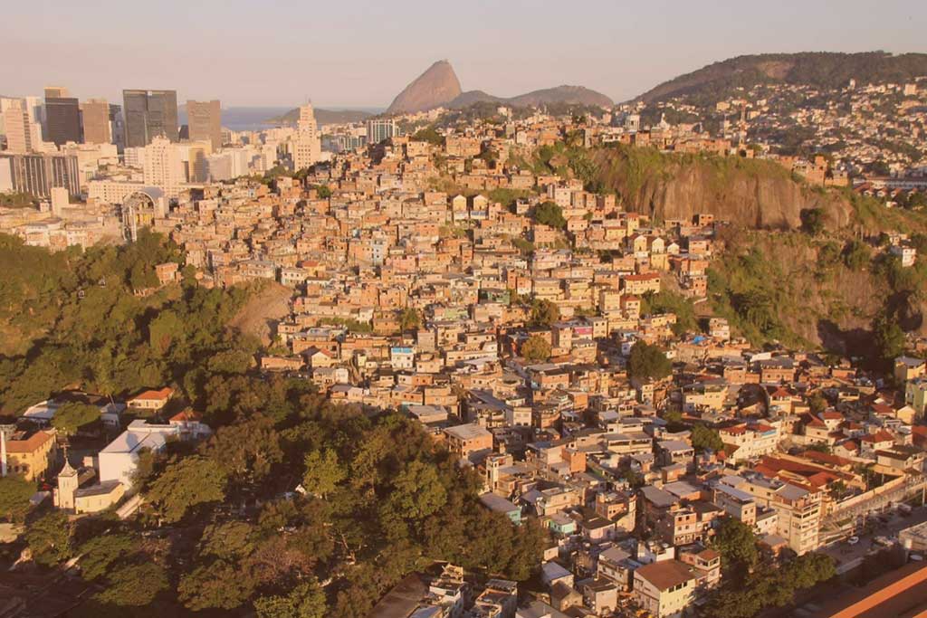 an aerial view of a shantytown on a steep hill side in Rio, Sugerloaf Mountain is visible in the distance 