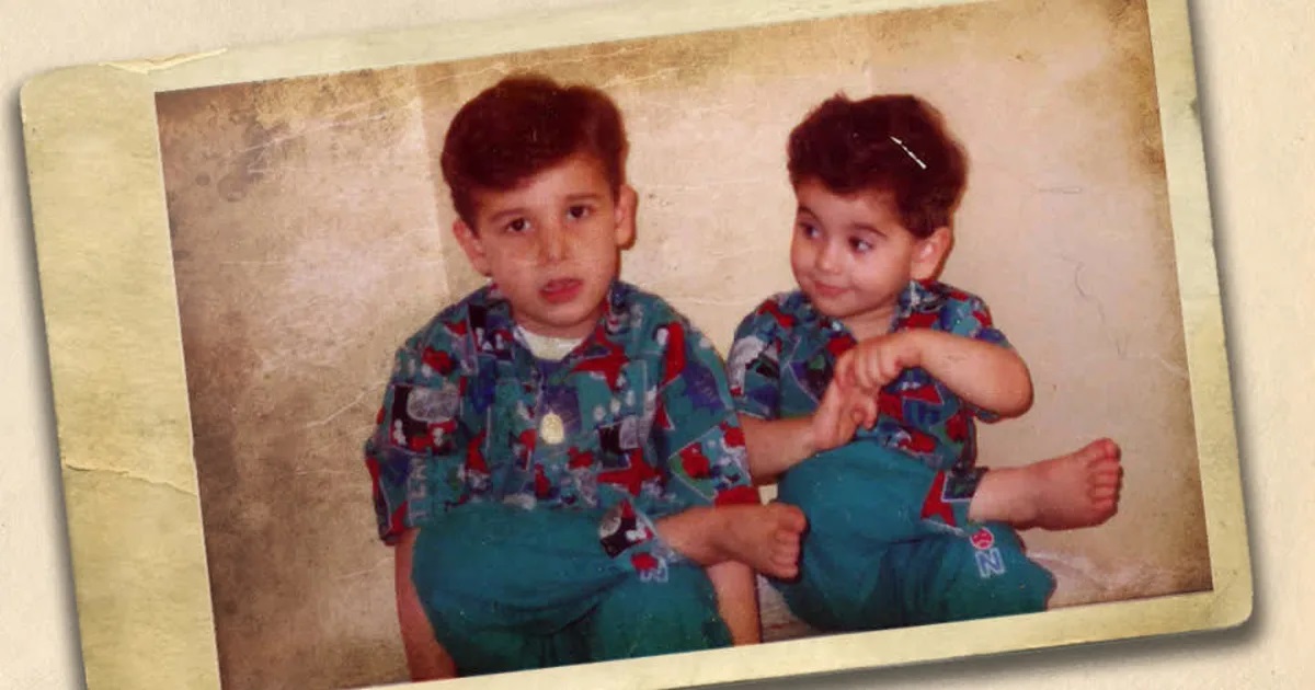 Two young brothers sit next to other, the younger looks to the elder.