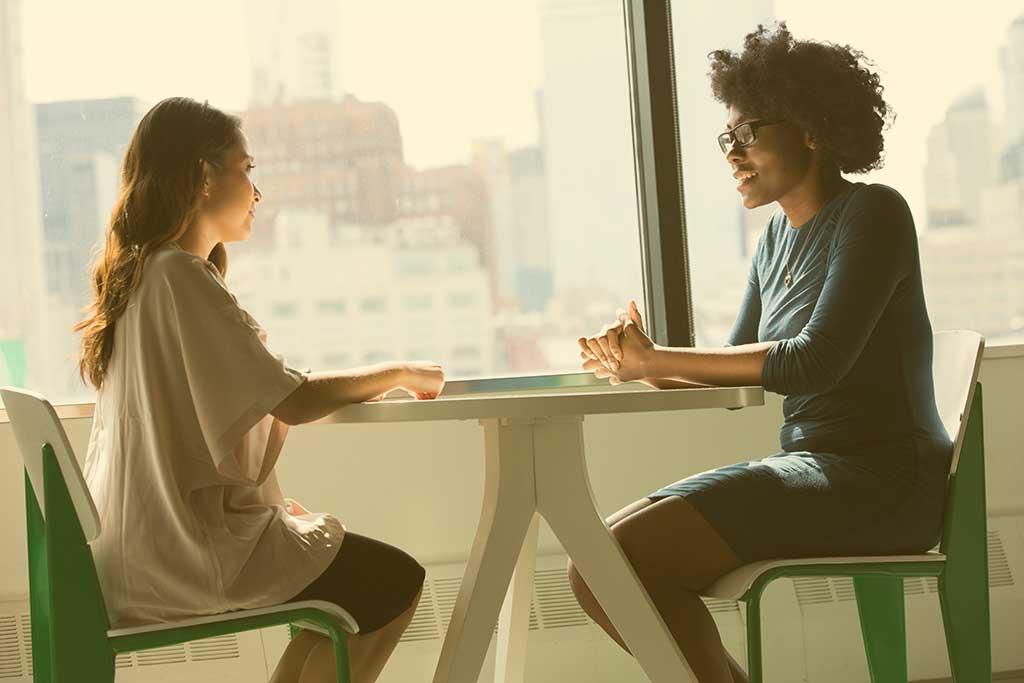 Two people sit at a table with their hands resting on top of it. One speaks to the other