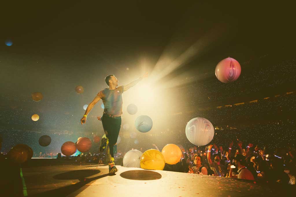 A singer struts a stage pointing to the spotlight as coloured orbs float down.