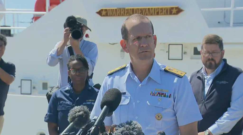 A Coast Guard officer gives a press conference while looking grim-faced. Others look on.