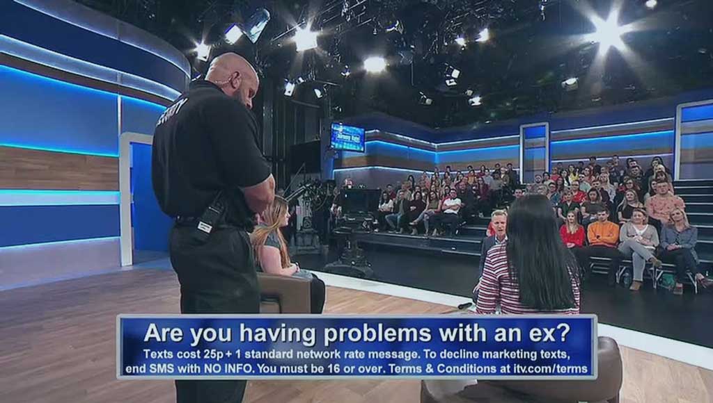 A view across a talk show TV set showing a security guard standing between two guest while the host talks to them. An audience looks on.