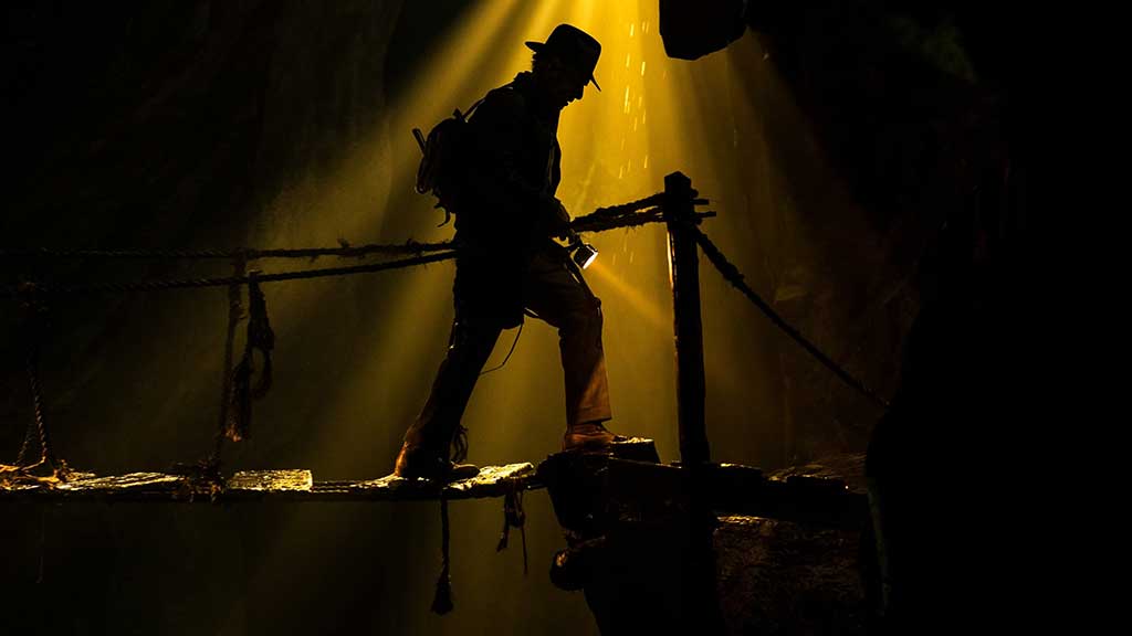 A silouhette of an adventurer, wearing a fedora hat, stepping gingerly along a rickety bridge.