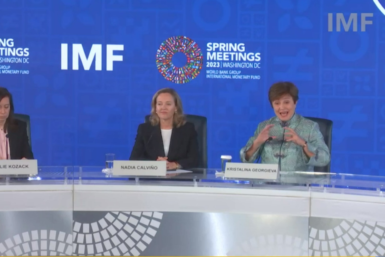 Two women sit behind a press conference desk against a backdrop, one listens as the other speaks and  gestures.