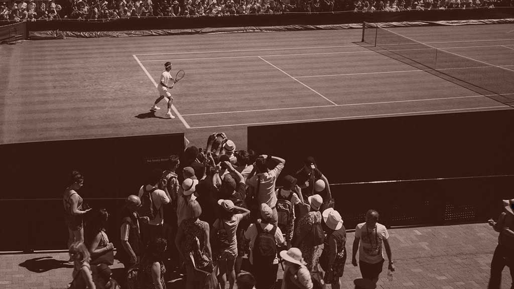 A tennis player stands ready to return a shot, while a phalanx of photographers crowd round a court-side opening to take a picture of him.