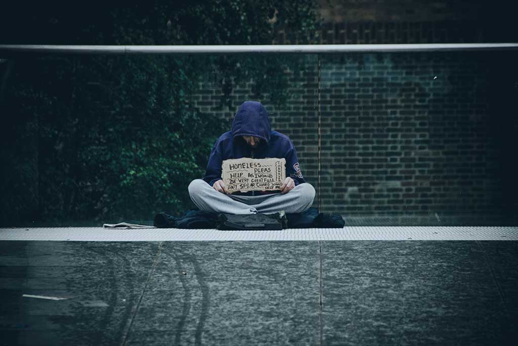 A beggar sits cross legged against a glass railing holding a sign.