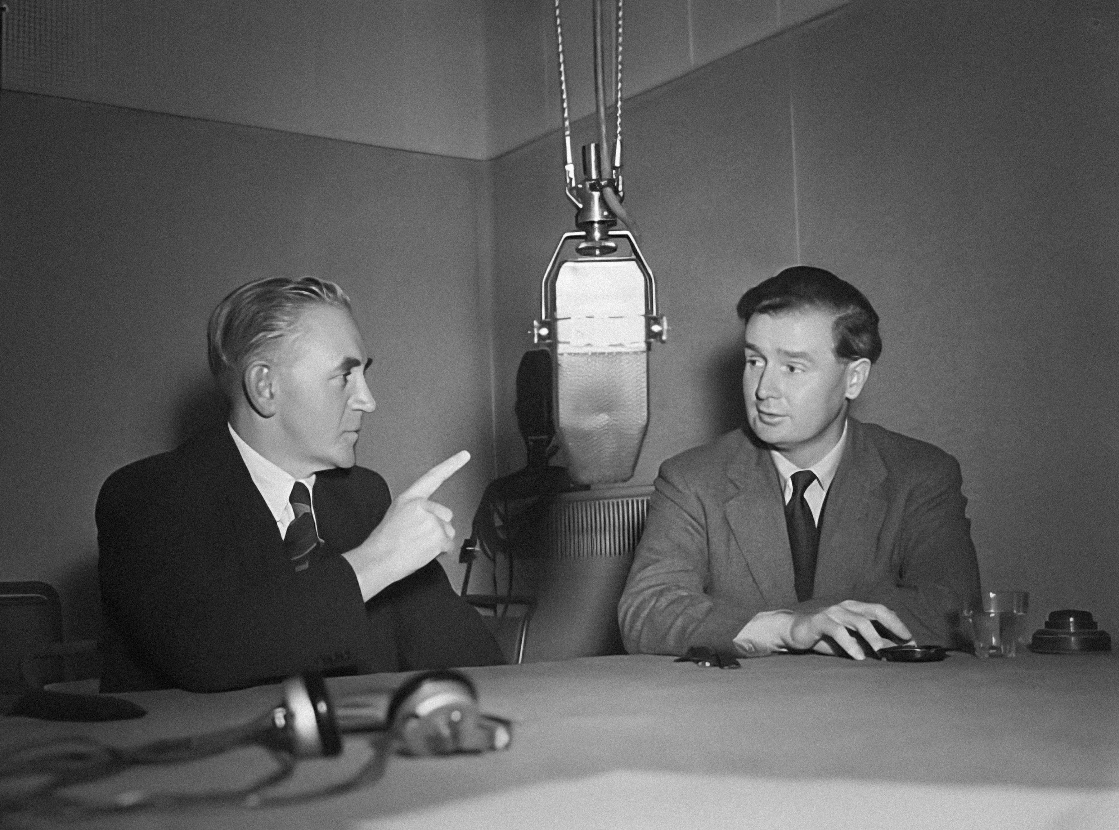 Two 1950's men un suits sit at a table dominated by a large hanging microphone. One points a raise hand and finger into the air. The other listens.