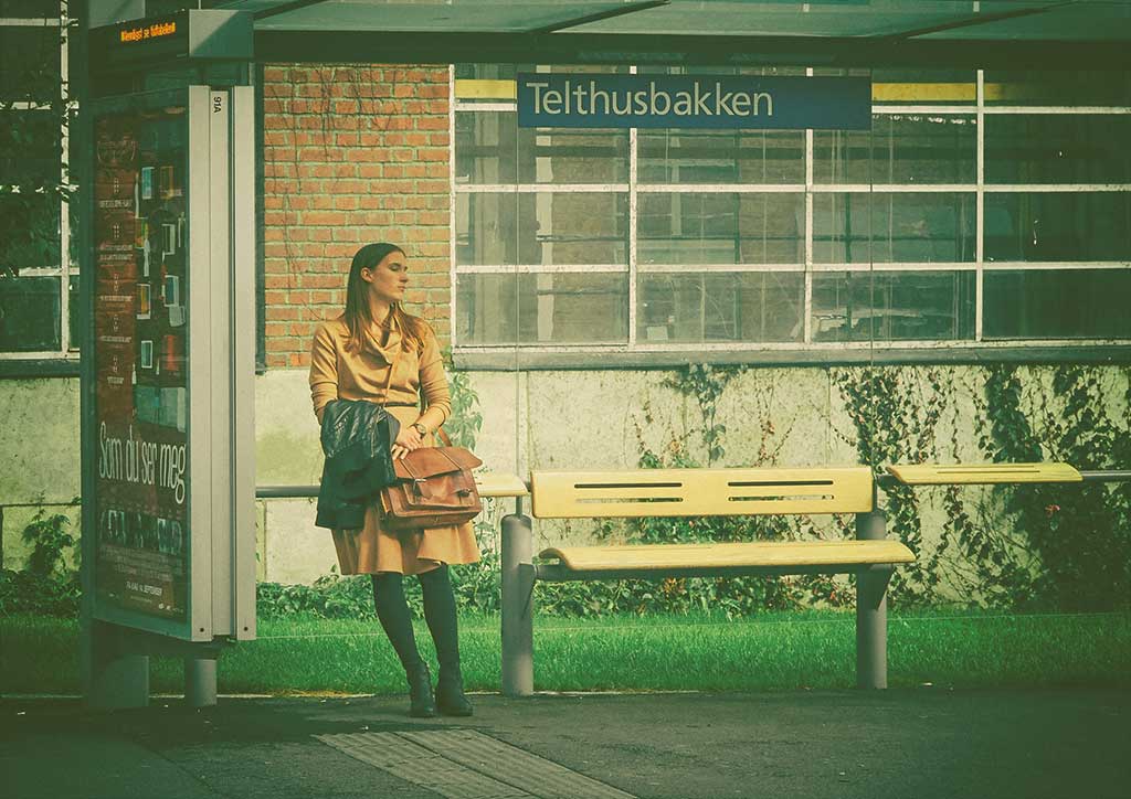 A woman leans against the glass of a bus shelter while waiting, she clasps a bag.