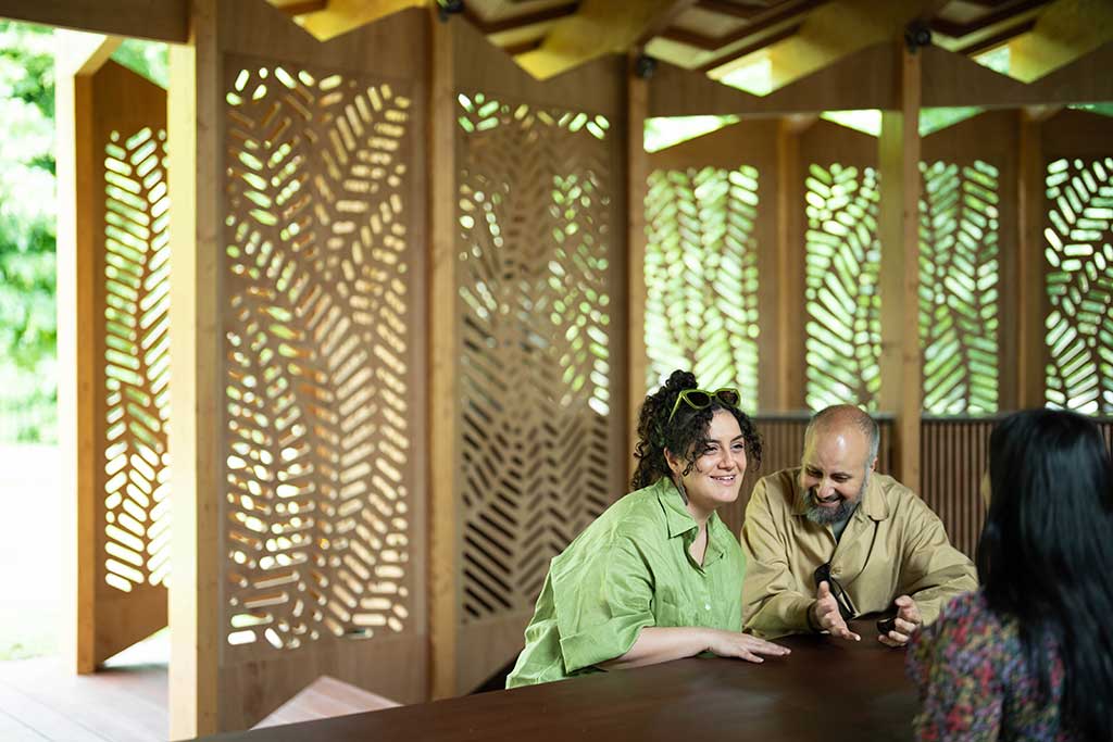 Around a table, against a backdrop of fret-cut wood, three people talk and listen to each other with great interest.