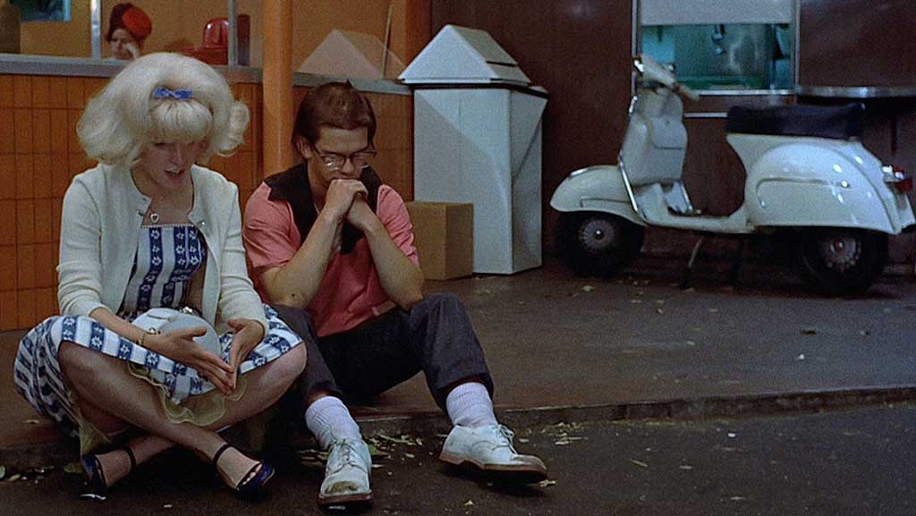 A teenage couple dressed in 1950s clothes sit on a kerb in deep conversation.