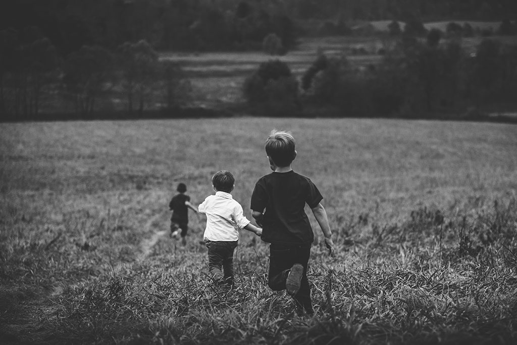 Three young children run away into the distance down across a field of long grass.